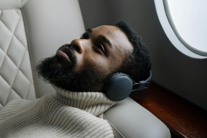 Man reclining on an airplane seat, wearing wireless headphones and a sweater, looking relaxed.