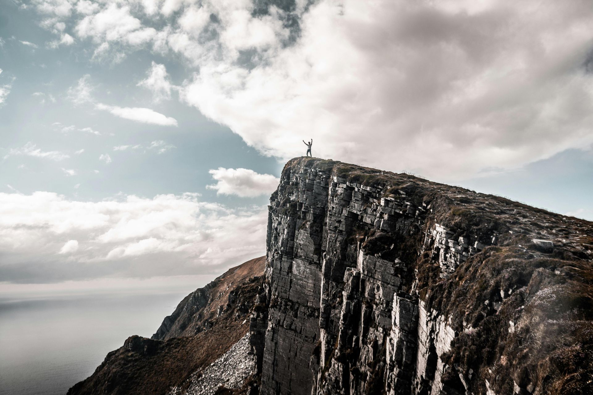 A lone hiker stands triumphantly on a rugged mountain cliff against a cloudy sky.